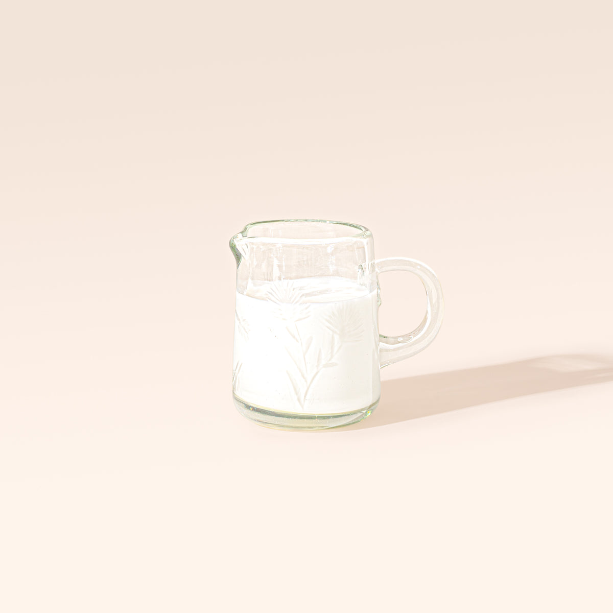 Clear glass creamer with etched floral designs, a rounded handle, and a pouring spout, filled with cream on a cream background.