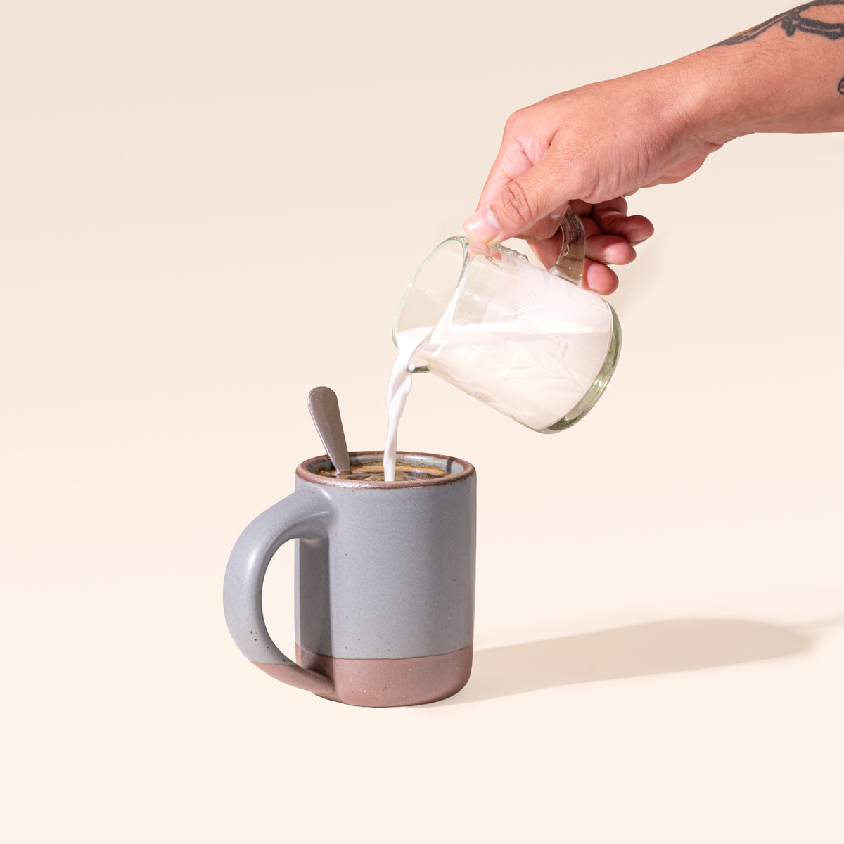 A ceramic mug in a blue-grey color with unglazed base with a hand pouring cream from a etched thistle glass creamer.