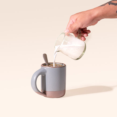 A ceramic mug in a blue-grey color with unglazed base with a hand pouring cream from a etched thistle glass creamer.