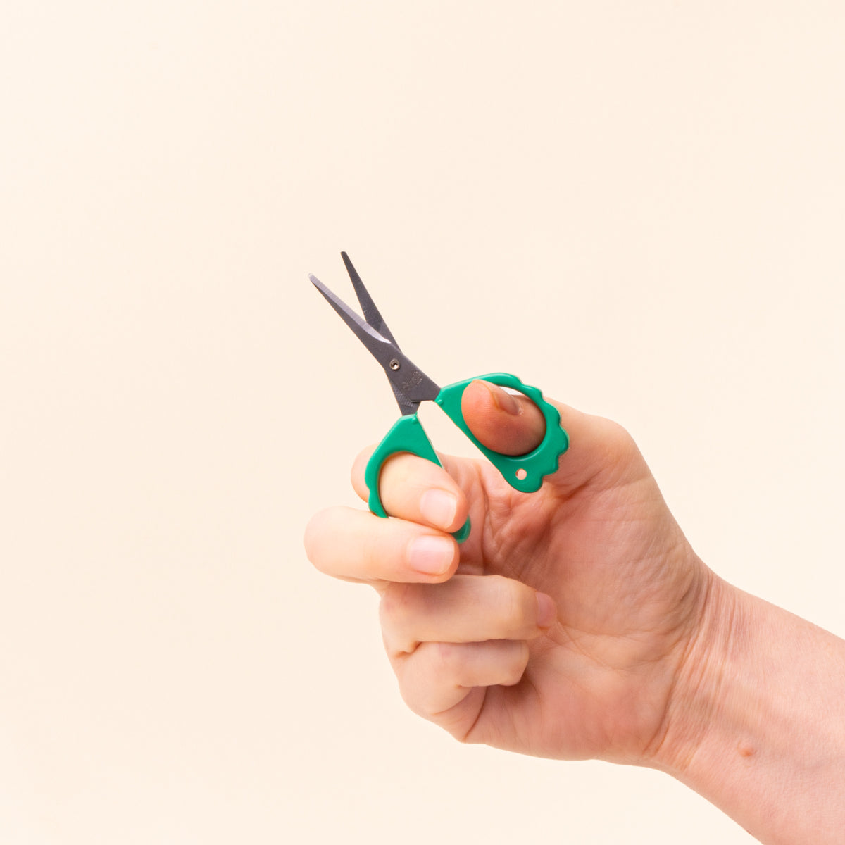 Hand holding small scissors with green scalloped handles and open black blades against a cream background.