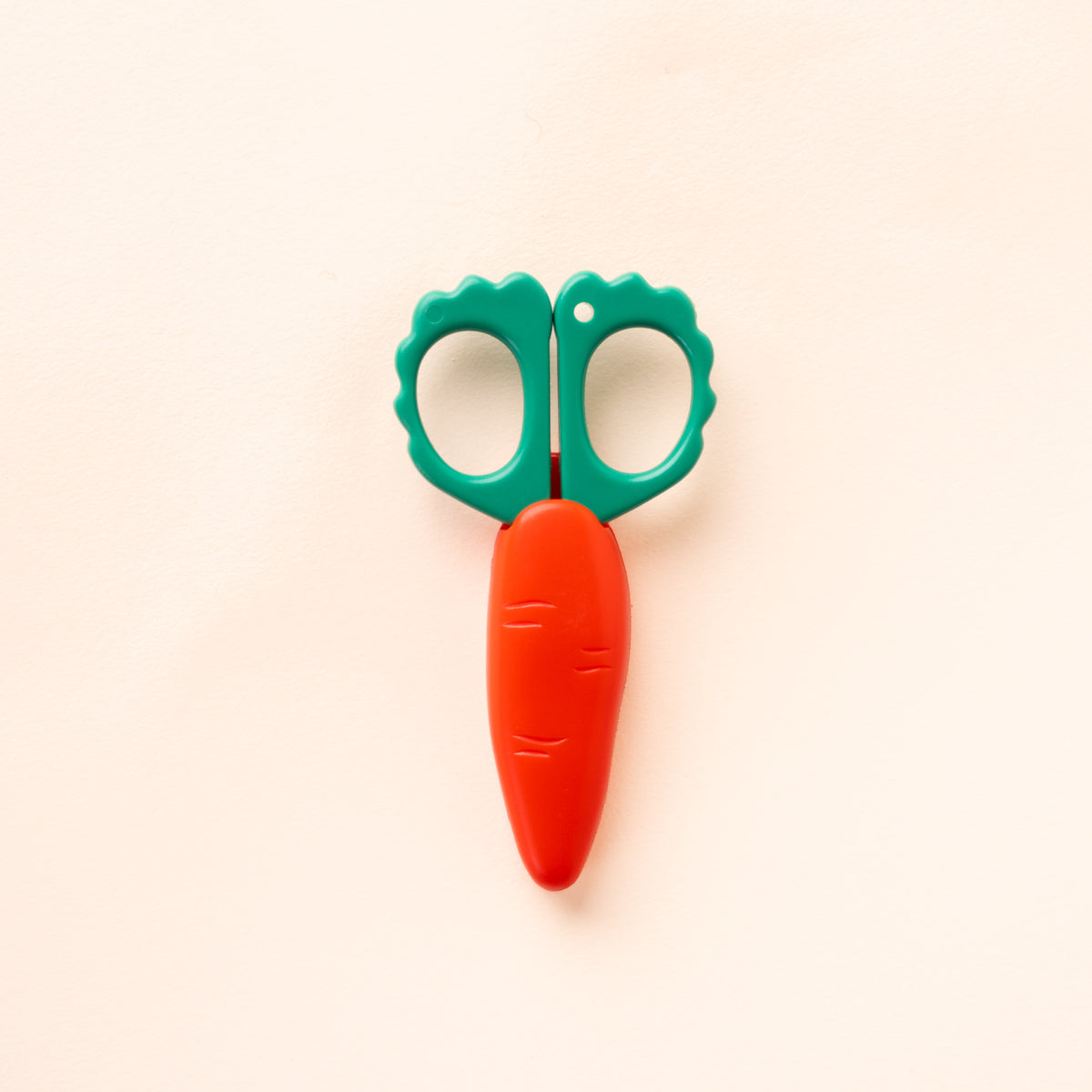 Carrot-shaped scissors with green scalloped handles and an orange protective blade cover, closed against a cream background.