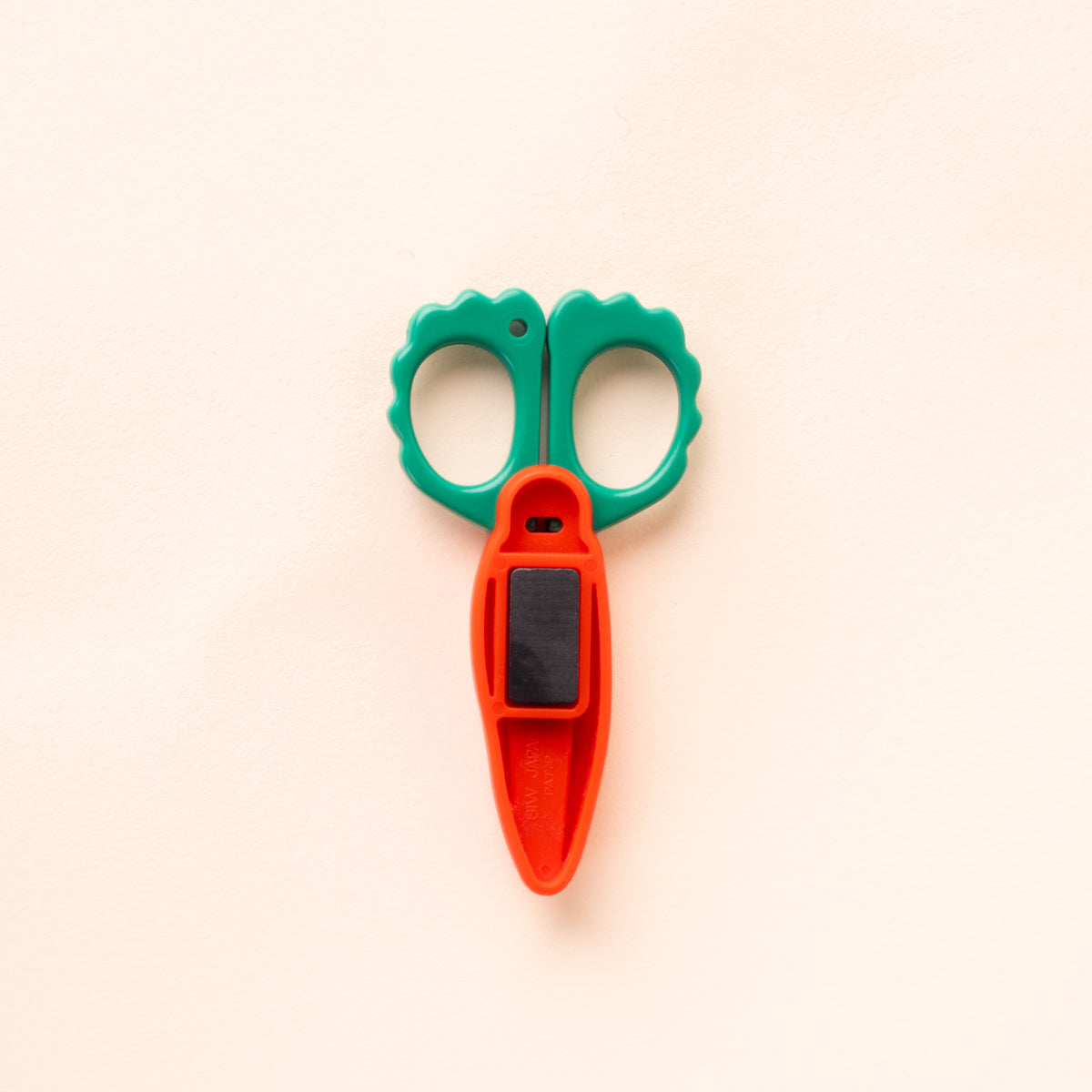 Back view of carrot-shaped scissors with green scalloped handles and an orange protective blade cover with a black magnetic strip.