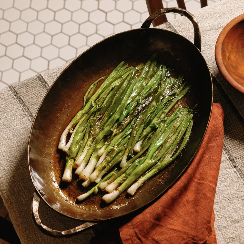 Roasted green onions in a black oval roasting pan on a linen-covered table with a wooden bowl and rust-colored napkin.