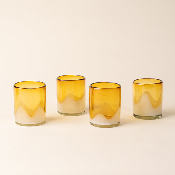 Four amber and clear handblown glass tumblers with organic wave design, casting a soft golden shadow on a beige backdrop.