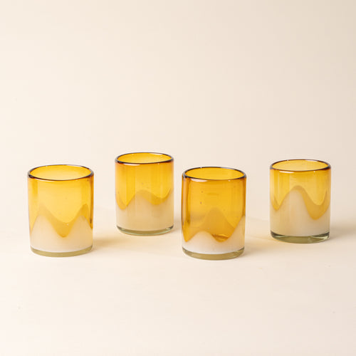 Four amber and clear handblown glass tumblers with organic wave design, casting a soft golden shadow on a beige backdrop.