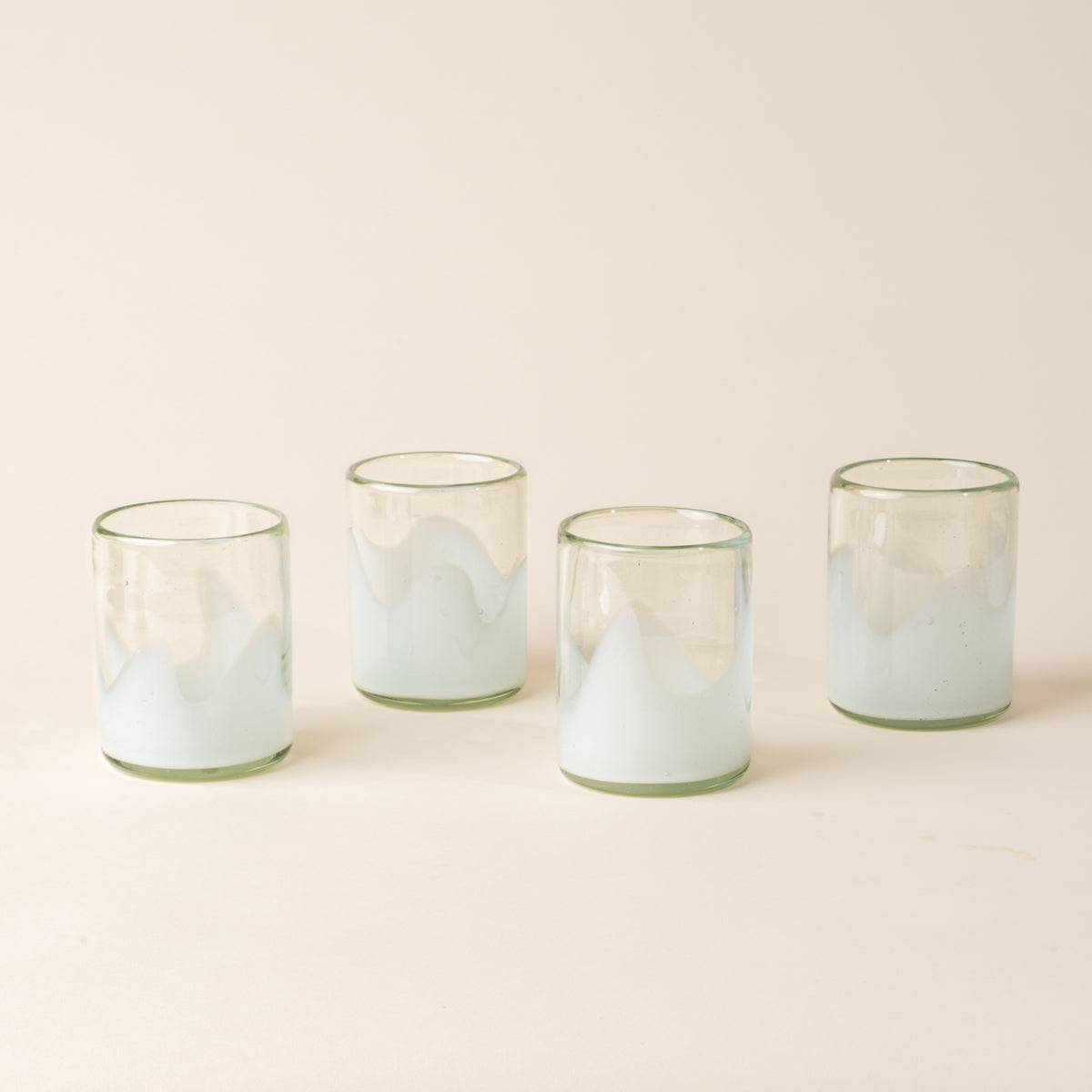 Four white and clear handblown glass tumblers with organic wave design, casting a soft golden shadow on a beige backdrop.