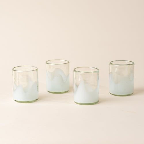 Four white and clear handblown glass tumblers with organic wave design, casting a soft golden shadow on a beige backdrop.