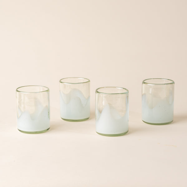 Four white and clear handblown glass tumblers with organic wave design, casting a soft golden shadow on a beige backdrop.