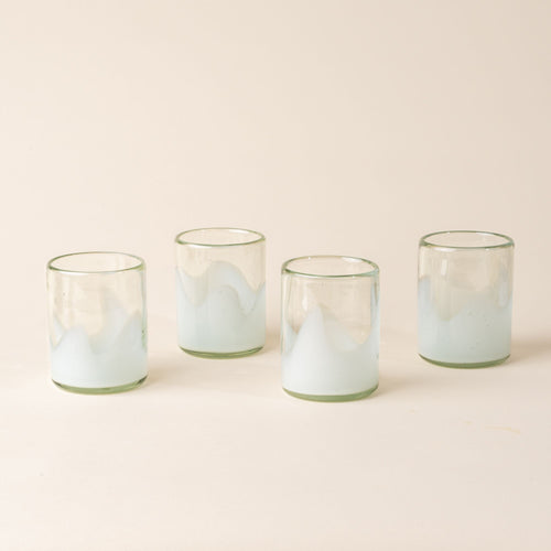 Four white and clear handblown glass tumblers with organic wave design, on a beige backdrop.