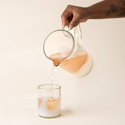 A hand holds a white and clear handblown glass pitcher with organic wave design with clear handle, and pours juice into a matching tumbler glass.