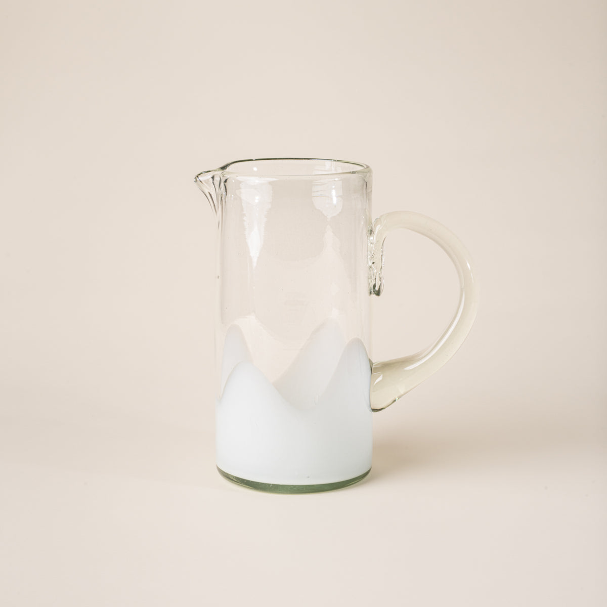 White and clear handblown glass pitcher with organic wave design with clear handle, casting a soft golden shadow on a beige backdrop.