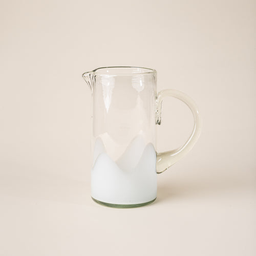 White and clear handblown glass pitcher with organic wave design with clear handle, casting a soft golden shadow on a beige backdrop.