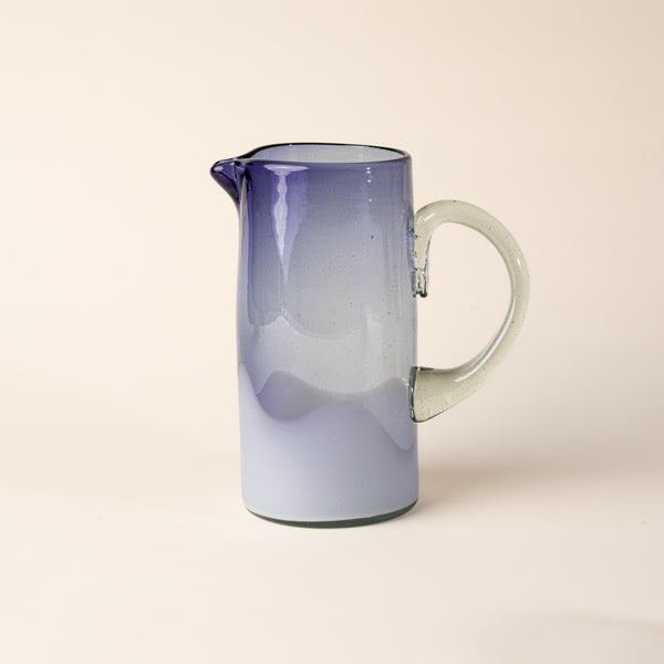 Violet and clear handblown glass pitcher with organic wave design with clear handle, casting a soft golden shadow on a beige backdrop.