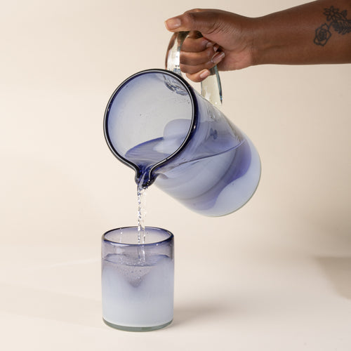 A hand holds a white and clear iris handblown glass pitcher with organic wave design with clear handle, and pours water into a matching tumbler glass.