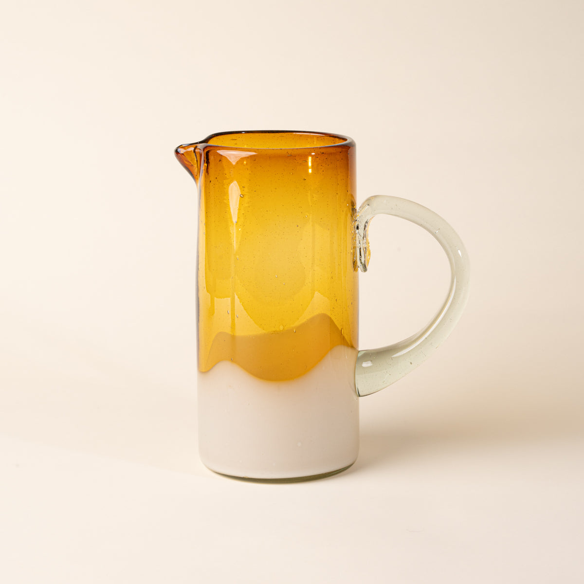 Amber and clear handblown glass pitcher with organic wave design with clear handle, casting a soft golden shadow on a beige backdrop.