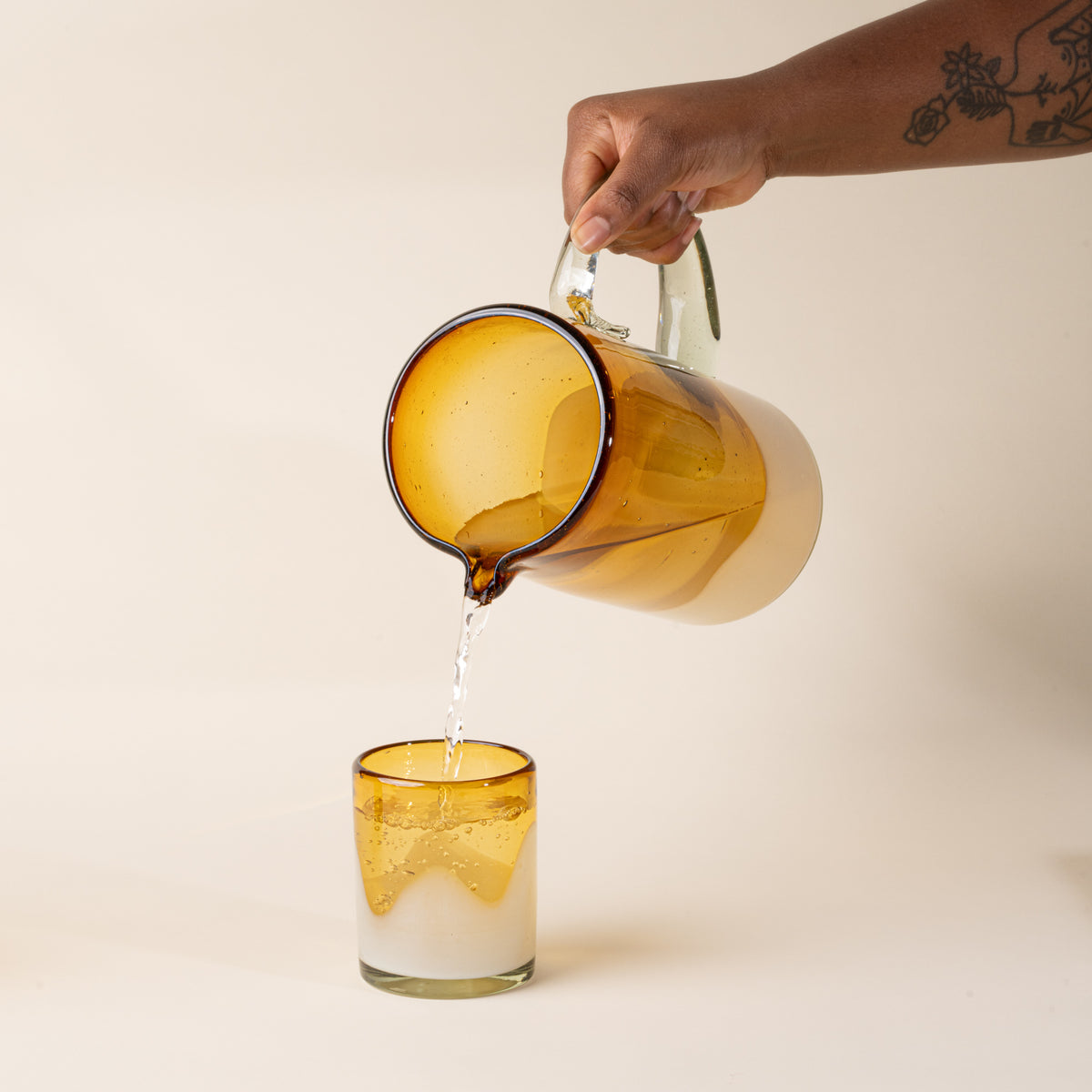 A hand holds a white and clear amber handblown glass pitcher with organic wave design with clear handle, and pours water into a matching tumbler glass.