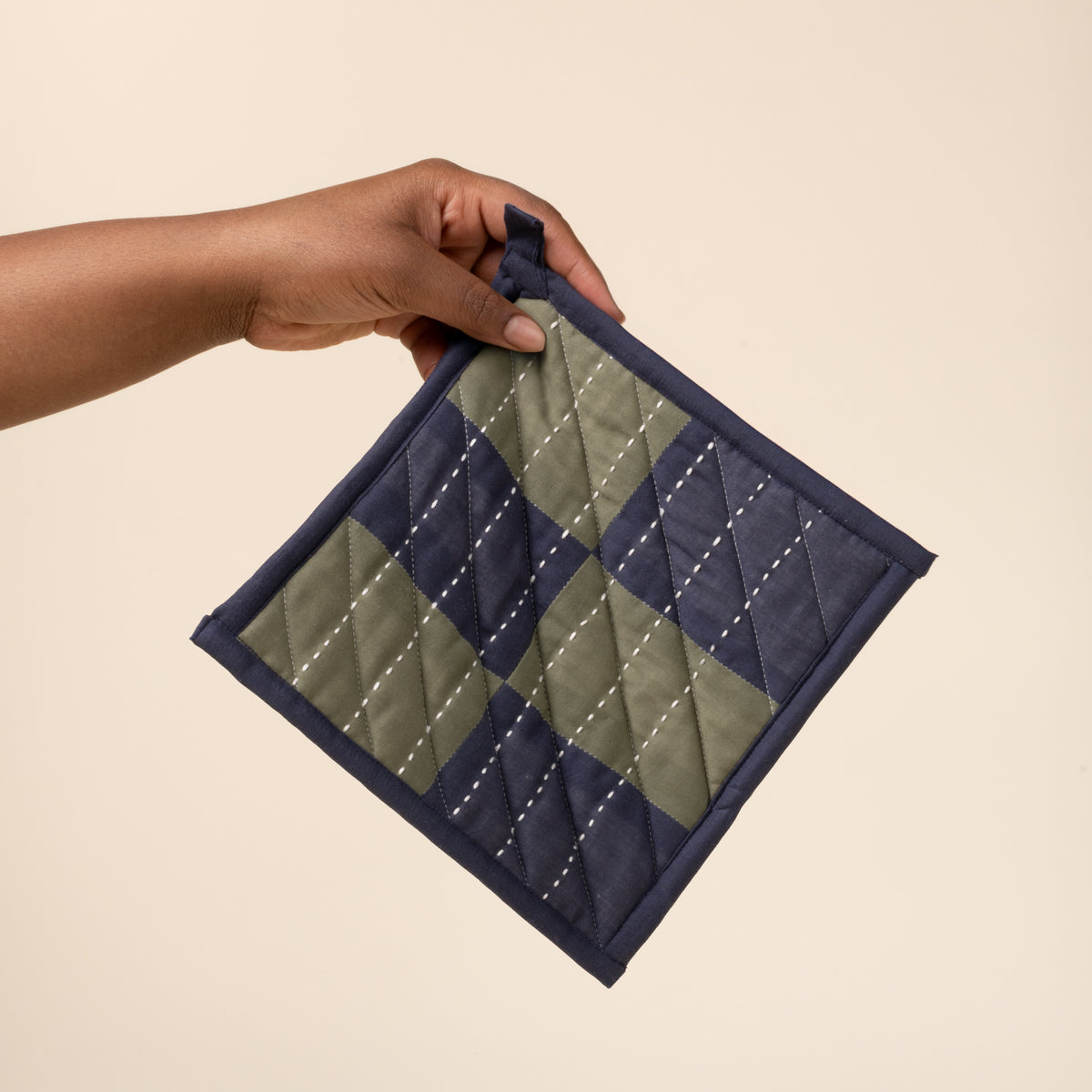 A hand holds a quilted pot holder with green and navy patchwork squares and white stitched lines, with hanging loop.