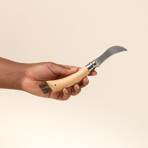 A hand holds a wood-handled folding mushroom knife with curved blade and built-in brush on handle end.