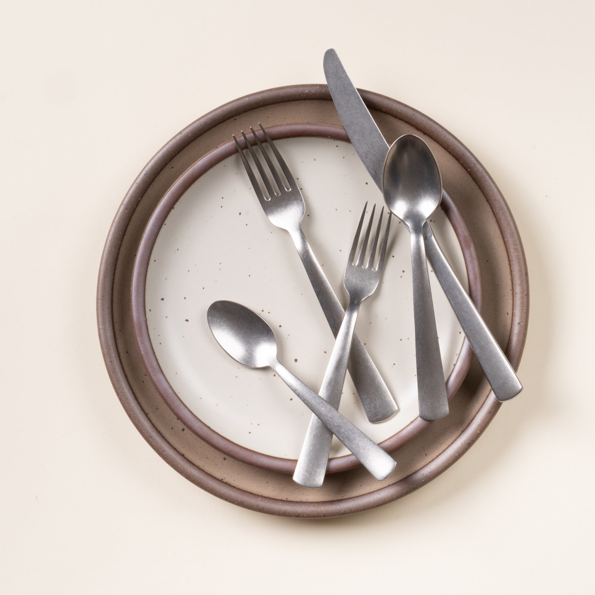 Five matte stainless steel utensils—two forks, knife, tablespoon, and teaspoon—resting on stacked speckled ceramic plates.