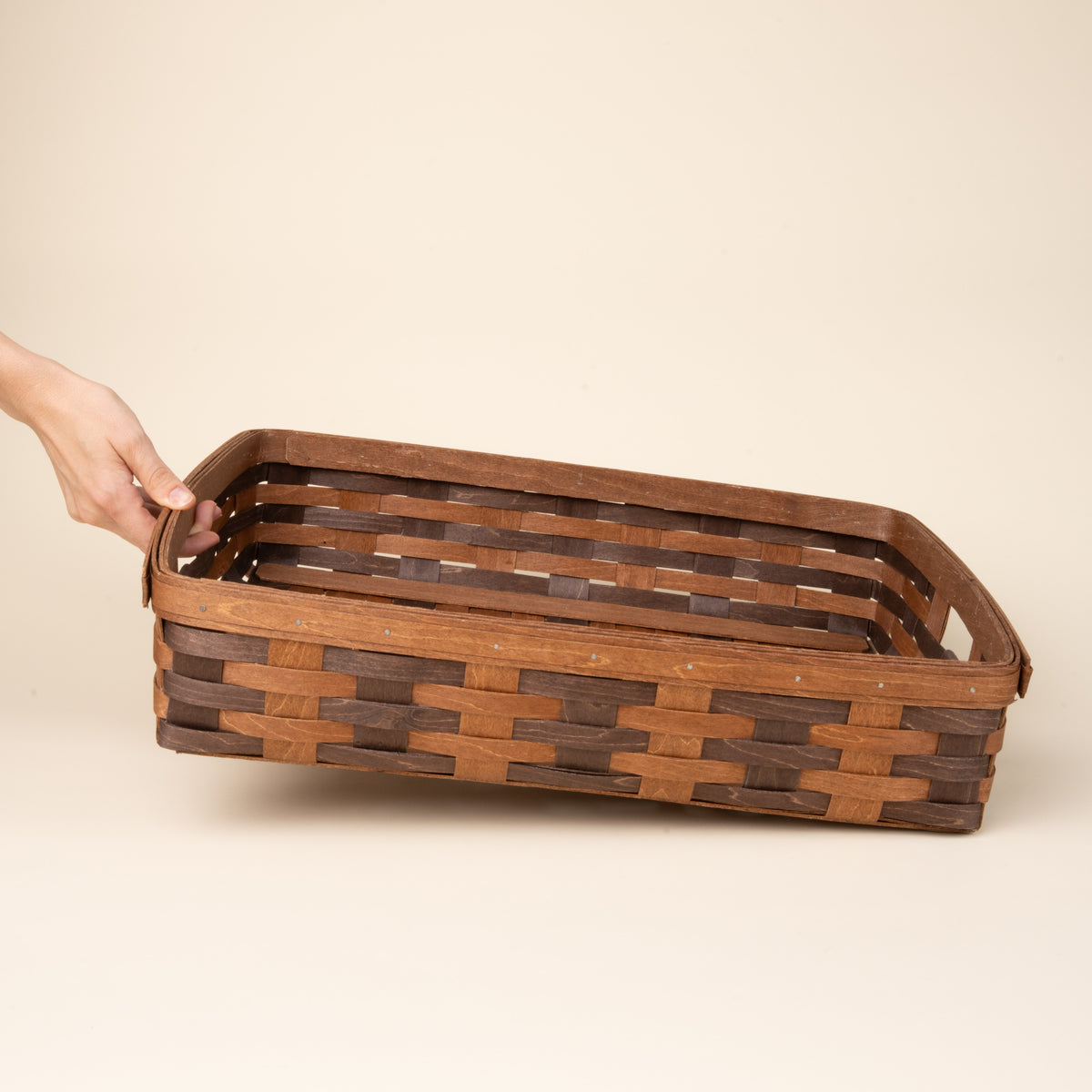 Hand holding one side of the rectangular woven wood basket in two-tone brown with an open top and reinforced rim, displayed against a neutral background.