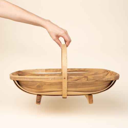 Hand holds a large oval wooden gathering trug basket with a center handle and small feet.