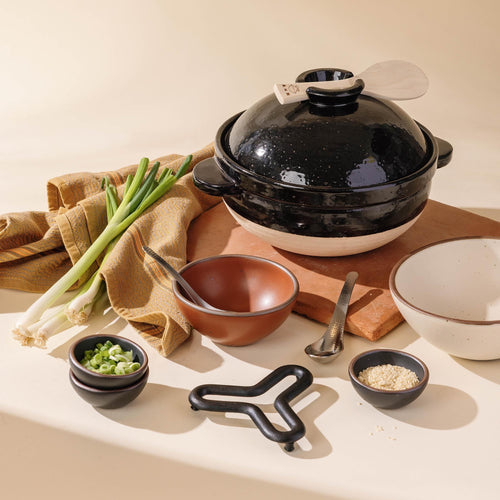 Black Donabe styled with fresh scallions, sesame, and serving pieces for a modern, elevated kitchen vignette.