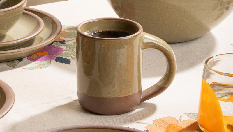 Ceramic mug with coffee, plate with pastry, bowl of oranges on a floral tablecloth.