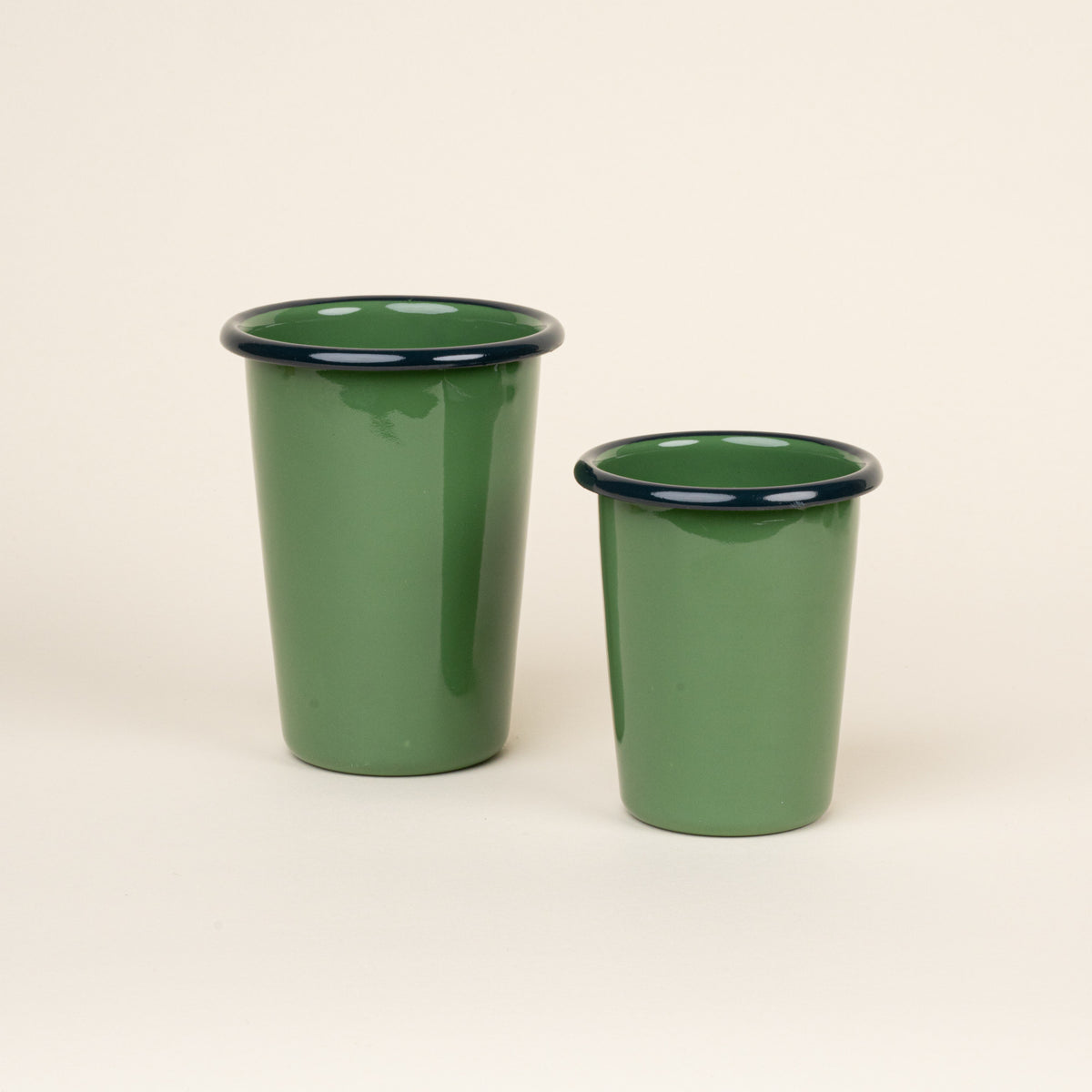 A short and tall green enamel tumbler with a dark rim, set against a soft beige background.