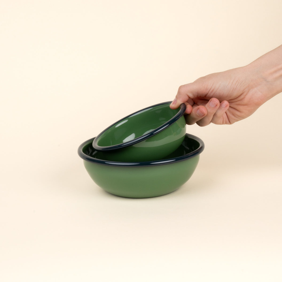 A hand holds a small enamel bowl to nest in a medium enamel bowl; both bowls are green with a dark rim.