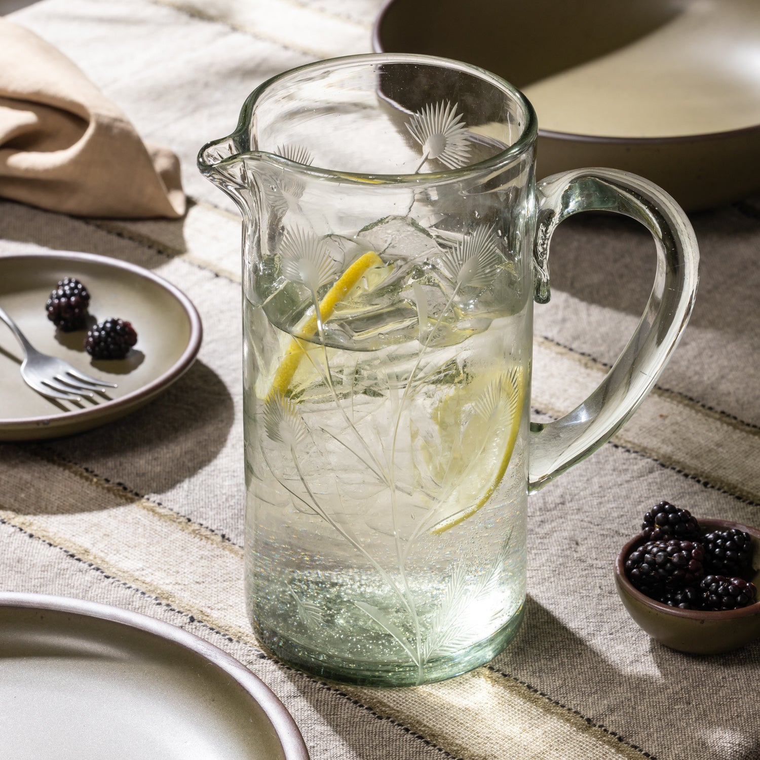 Glass pitcher with etched botanical pattern filled with ice water and lemon slices on a table with bowls of blackberries and ceramic dinnerware.