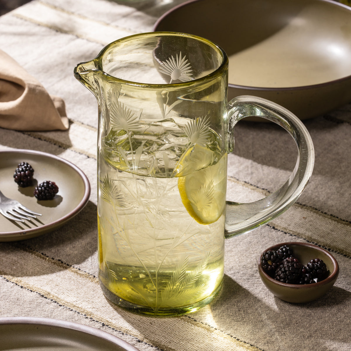 Olive glass pitcher with etched botanical pattern filled with ice water and lemon slices on a table with bowls of blackberries and ceramic dinnerware.