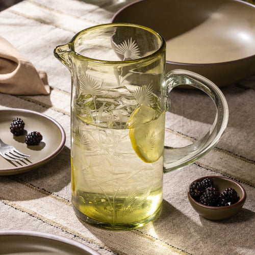 Olive glass pitcher with etched botanical pattern filled with ice water and lemon slices on a table with bowls of blackberries and ceramic dinnerware.