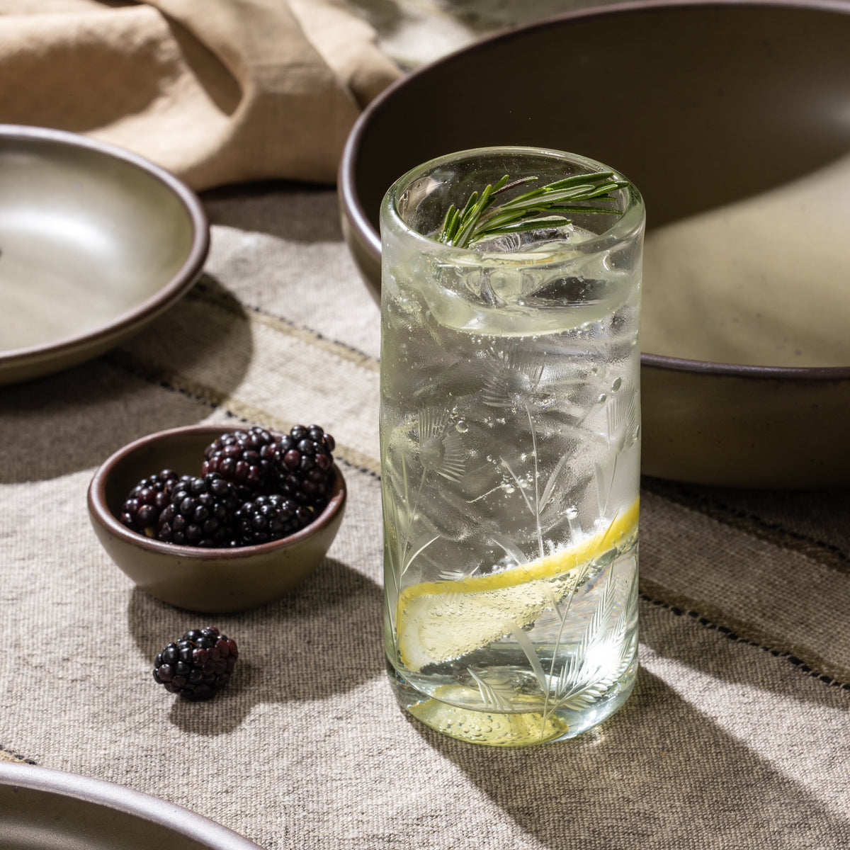Etched glass tumbler filled with ice, lemon slices, and a rosemary sprig beside a bowl of blackberries and ceramic dinnerware.