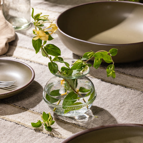 Round clear glass bud vase holding flowering honeysuckle stems on a table beside ceramic dinnerware.