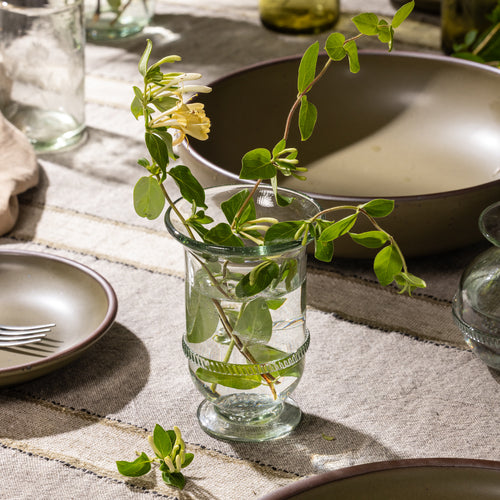 Tall clear glass bud vase holding flowering honeysuckle stems on a table beside ceramic dinnerware.