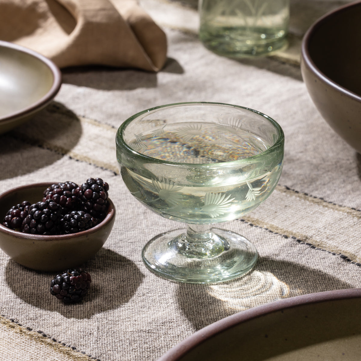 Etched glass coupe filled with a clear beverage beside a small bowl of blackberries on a table with ceramic dinnerware.
