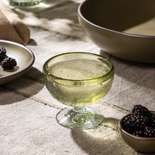Olive etched glass coupe filled with a clear beverage beside a small bowl of blackberries on a table with ceramic dinnerware.