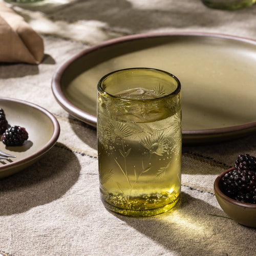 Olive etched glass tumbler filled with ice and a clear beverage beside a small bowl of blackberries and ceramic dinnerware.