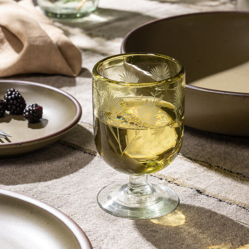 Olive etched stemmed glass filled with a clear beverage beside a small bowl of blackberries and ceramic dinnerware.