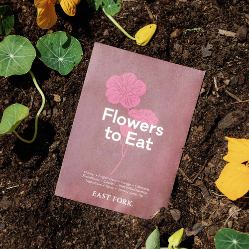 Flowers to Eat seed packet with pink floral illustration outside in dirt with greenery.