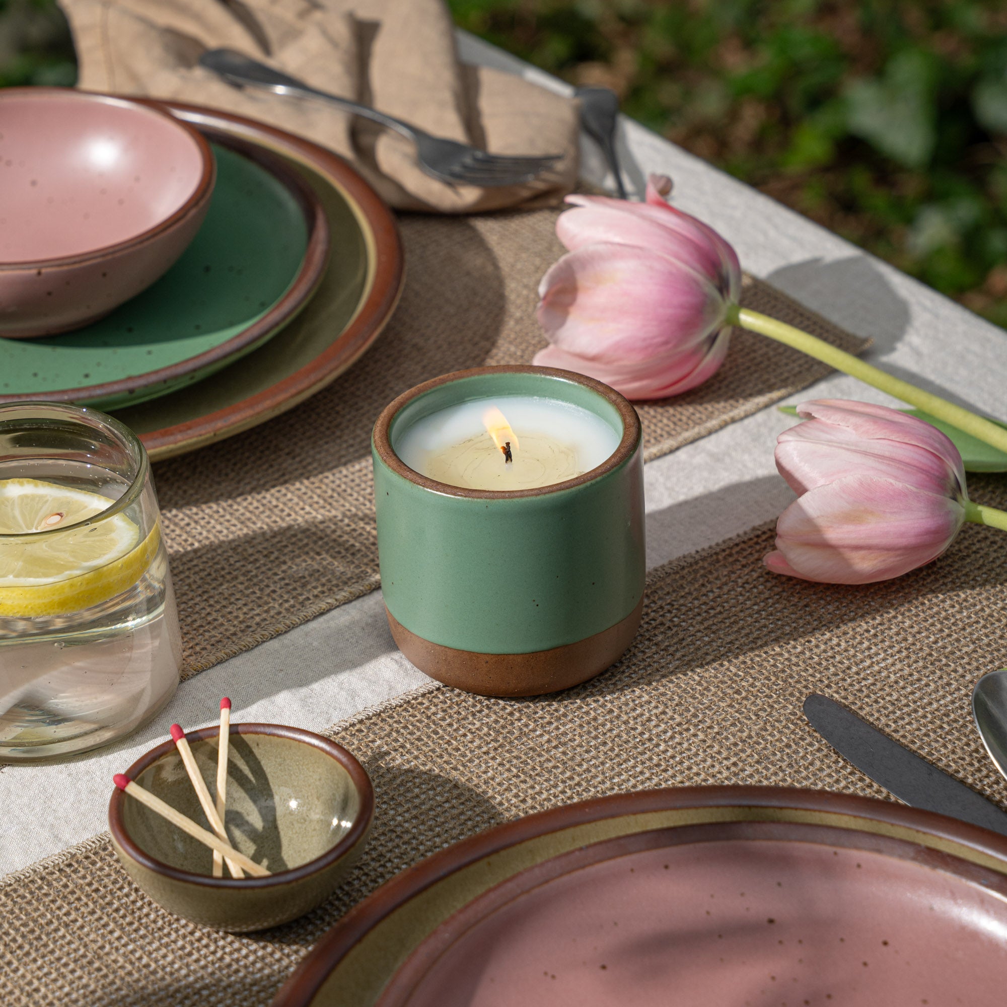 Lit green ceramic candle on a table setting with plates, glass of lemon water, matches, and pink tulips outdoors.