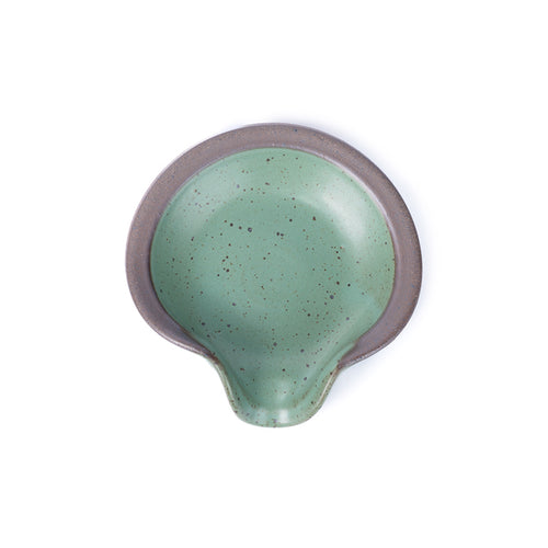 A circular ceramic spoon rest in a green color featuring iron speckles and unglazed ridge rim