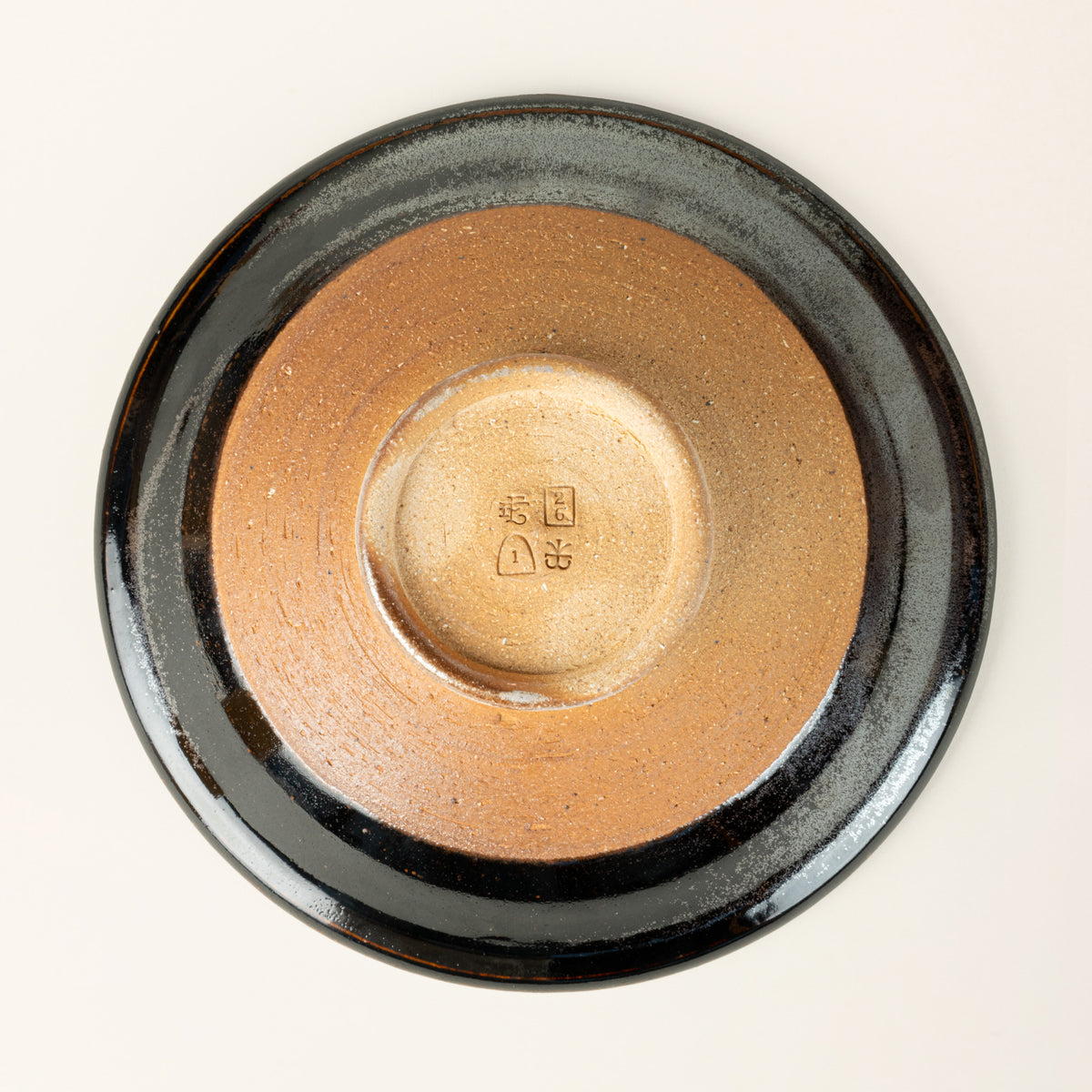 The bottom of a small handmade ceramic bowl with a wood fired earthy muddy glaze and an unglazed base, showing 4 stamps.