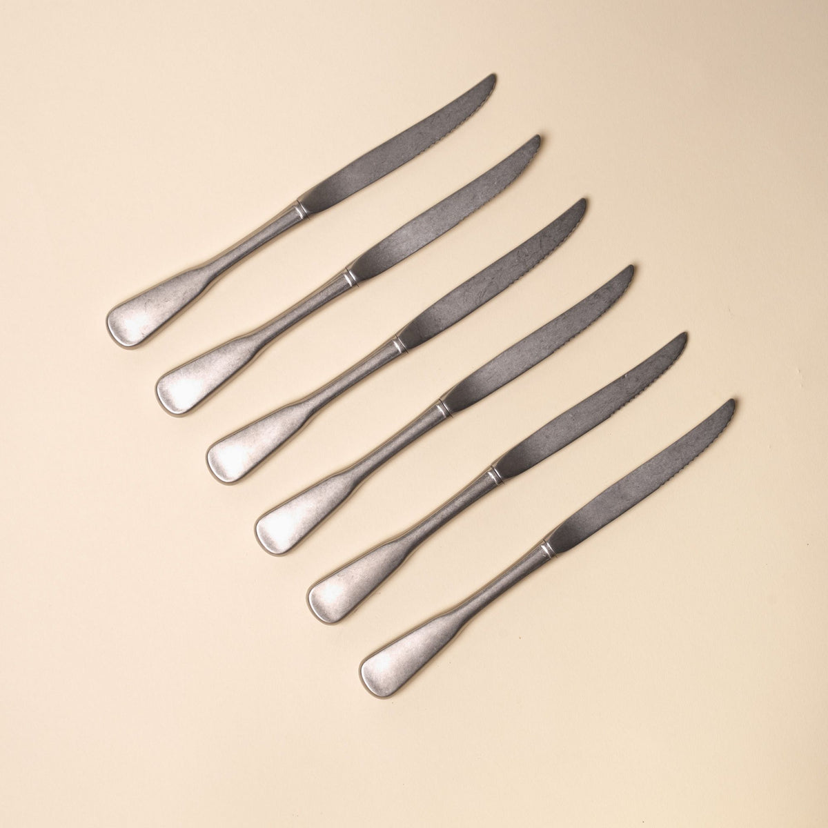 Six matte stainless steel dinner steak knives with rounded handles arranged diagonally on a neutral beige background.