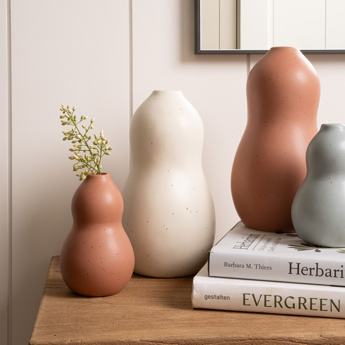 Four curvy vases in terracotta, cream, and blue-grey tones arranged on a wooden surface, with two vases resting on stacked books.