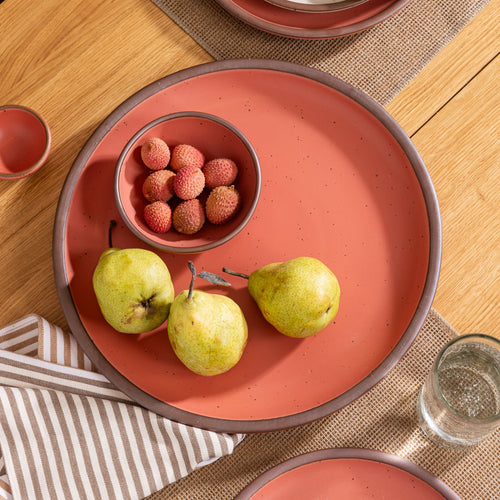 Watermelon pink serving platter with three green pears and a small bowl of lychees, set on a wood table with a striped napkin.