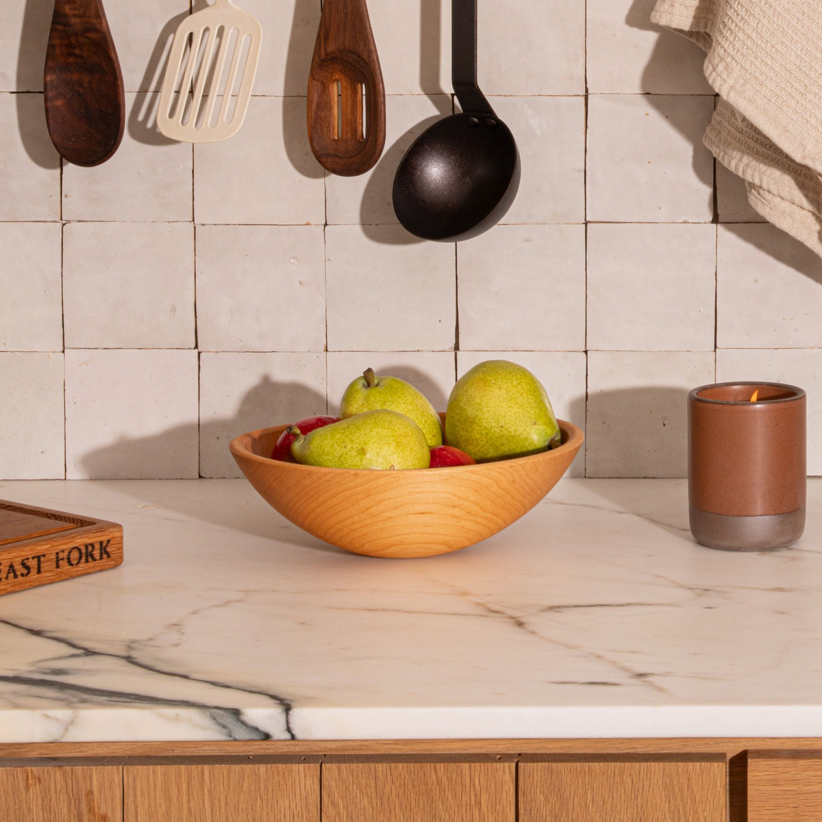 Wooden bowl with pears and apples on marble counter beside a lit brown candle, with utensils hanging on tiled wall.