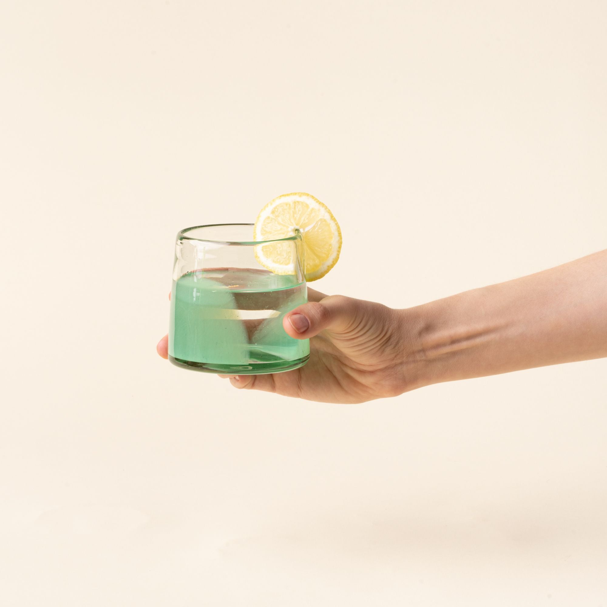 A hand holds out a short clear glasses with pastel green arch base filled with water and a lemon slice.