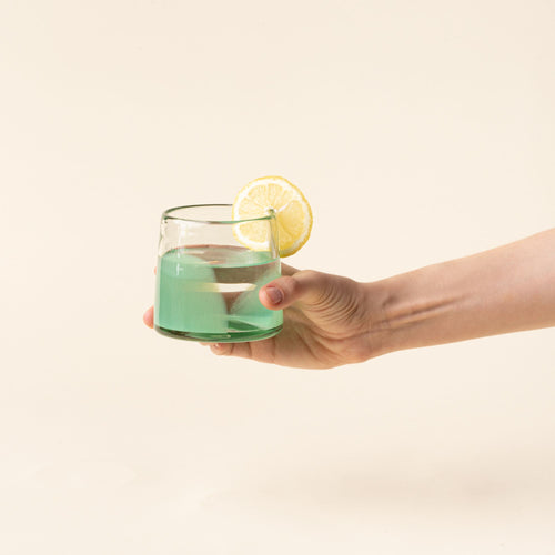 A hand holds out a short clear glasses with pastel green arch base filled with water and a lemon slice.