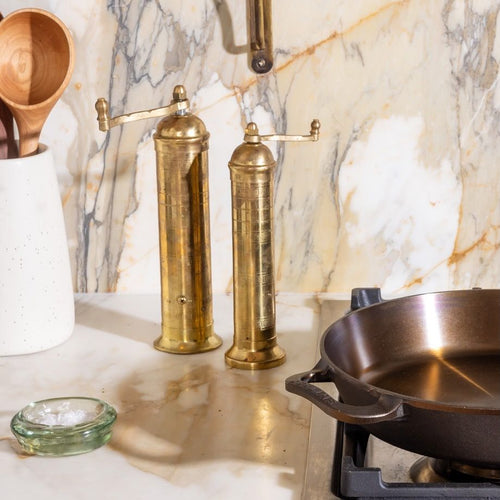Two tall brass salt and pepper mills on a marble counter beside a cast-iron skillet and wooden utensils.